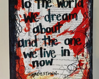 May include: Mixed media artwork with the text "To the world we dream about and the one we live in now" in black and teal lettering. Red paint strokes and scribbles overlay a background of sheet music and text. The word "HADESTOWN" is at the bottom.