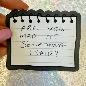 May include: A rectangular sticker shaped like a spiral-bound notepad page with a black border. The white paper features horizontal gray lines and the handwritten black text: ARE YOU MAD AT SOMETHING I SAID? The design includes black spiral loops at the top.