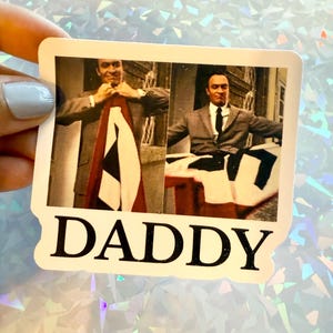 May include: A sticker featuring a vintage black and white image of a man in a suit. The word "DADDY" is printed in bold, black letters at the bottom of the sticker. The background is a holographic, iridescent surface.