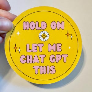 May include: A round yellow sticker with the text "HOLD ON LET ME CHAT GPT THIS" in pink bubble letters. A white daisy with a smiling face is in the center. The sticker has a retro design with starburst accents.