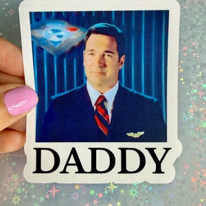 May include: A rectangular sticker featuring a portrait of a man in a dark blue suit, white shirt, and red and blue striped tie. He has a small gold pin on his lapel. Below the image, the word DADDY is printed in large, bold, black serif font.