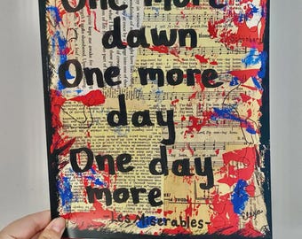 May include: A rectangular art print with the words "One more dawn, One more day, One day more" in black, over a background of sheet music and red and blue paint splatters. The text "Les Miserables" is at the bottom.