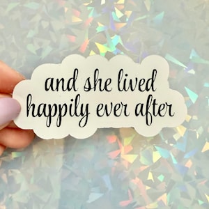 May include: A white sticker in a cloud shape with the phrase "and she lived happily ever after" in black cursive script. The sticker is held against a shimmering, iridescent background.