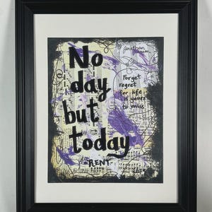 May include: Framed artwork with the words "No day but today" in black lettering. The background features sheet music, purple paint strokes, and handwritten script. The black frame surrounds a white mat.