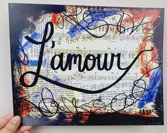 May include: Artwork featuring the word "L'amour" in elegant script over a background of sheet music and abstract brushstrokes in red, white, and blue. The artwork has a vintage aesthetic.