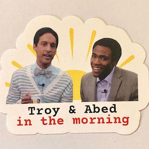 May include: A sticker featuring a cartoon sun with two men in front of it. The men are smiling and holding microphones. The text "Troy & Abed in the morning" is below the image.
