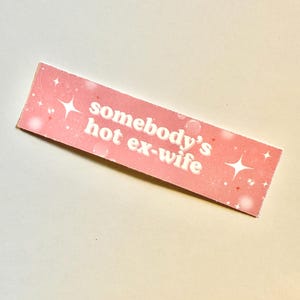 May include: A rectangular pink sticker with the text "somebody's hot ex-wife" in white. The sticker has a sparkly design with white stars and dots. The sticker is a novelty item.