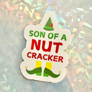 May include: A white sticker with the text "SON OF A NUTCRACKER" in green and red. The sticker features a green elf hat with a red accent and green elf shoes with yellow legs.