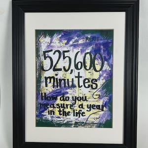 May include: Framed artwork with a black frame and white matting. The artwork features the text "525,600 minutes" and "How do you measure a year in the life" in black lettering. The background has green, purple, and yellow brushstrokes.
