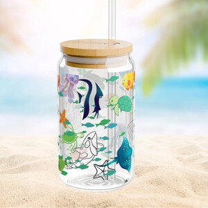 May include: A clear glass tumbler with a bamboo lid and a glass straw. The exterior features a colorful print of various marine life, including a jellyfish, angelfish, sea turtle, starfish, whale, and stingray, surrounded by small green fish.