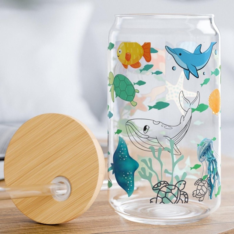 May include: A clear glass tumbler with a bamboo lid and a glass straw, featuring a colorful underwater print of a yellow fish, blue dolphin, green sea turtle, whale, stingray, jellyfish, and orange seashell scattered across the surface.