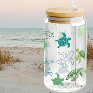 May include: A clear glass tumbler with a bamboo lid and a glass straw, featuring a repeating pattern of sea turtles in various shades of green and blue. The design includes both solid-colored and outlined turtle illustrations on the glass surface.