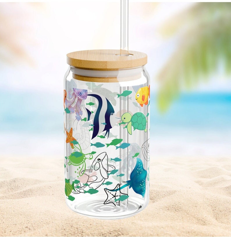 May include: A clear glass tumbler with a bamboo lid and a glass straw. The exterior features a colorful print of various marine life, including a jellyfish, angelfish, sea turtle, starfish, whale, and stingray, surrounded by small green fish.