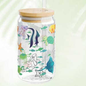 May include: A clear glass tumbler with a bamboo lid and a glass straw. The exterior features a colorful pattern of various sea creatures including a jellyfish, angelfish, sea turtle, starfish, whale, and stingray, interspersed with small green fish.