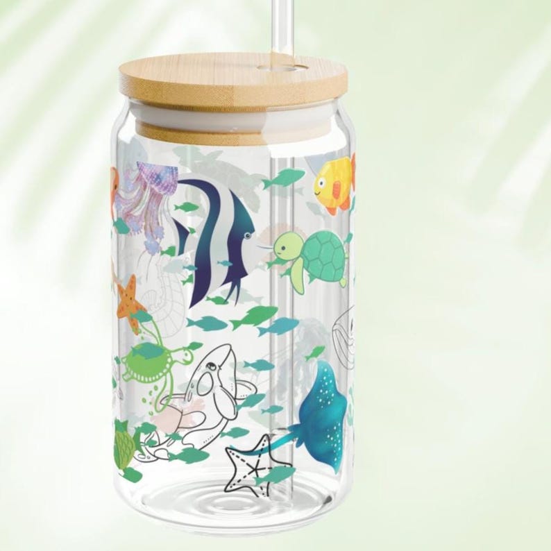 May include: A clear glass tumbler with a bamboo lid and a glass straw. The exterior features a colorful pattern of various sea creatures including a jellyfish, angelfish, sea turtle, starfish, whale, and stingray, interspersed with small green fish.