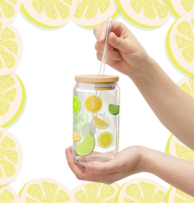 May include: A clear glass tumbler with a bamboo lid and a glass straw, featuring a colorful pattern of yellow lemon and green lime slices printed on the exterior. The cylindrical glass container is designed for beverages and includes a straw hole.