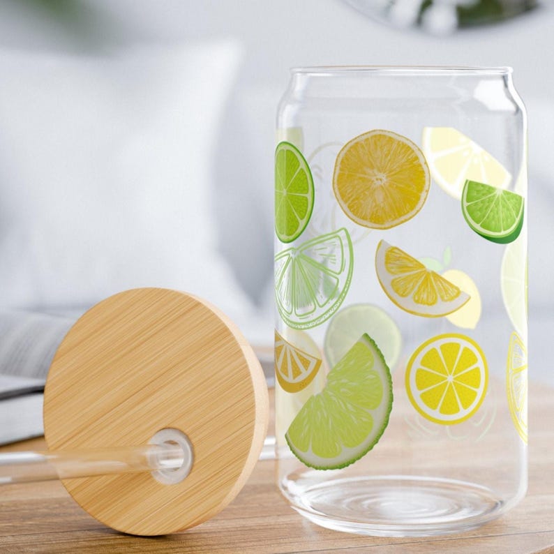 May include: A clear glass tumbler with a cylindrical shape featuring a colorful pattern of yellow lemon and green lime slices. The set includes a round bamboo lid with a central hole and a clear glass straw, designed for cold beverages.