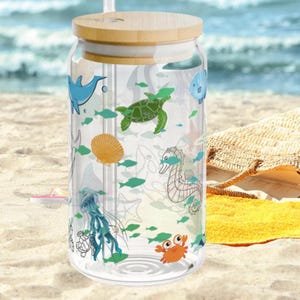 May include: A clear glass tumbler with a bamboo lid and a glass straw, featuring a colorful wrap-around print of various sea creatures including a blue dolphin, green sea turtle, orange crab, seahorse, jellyfish, and seashells.