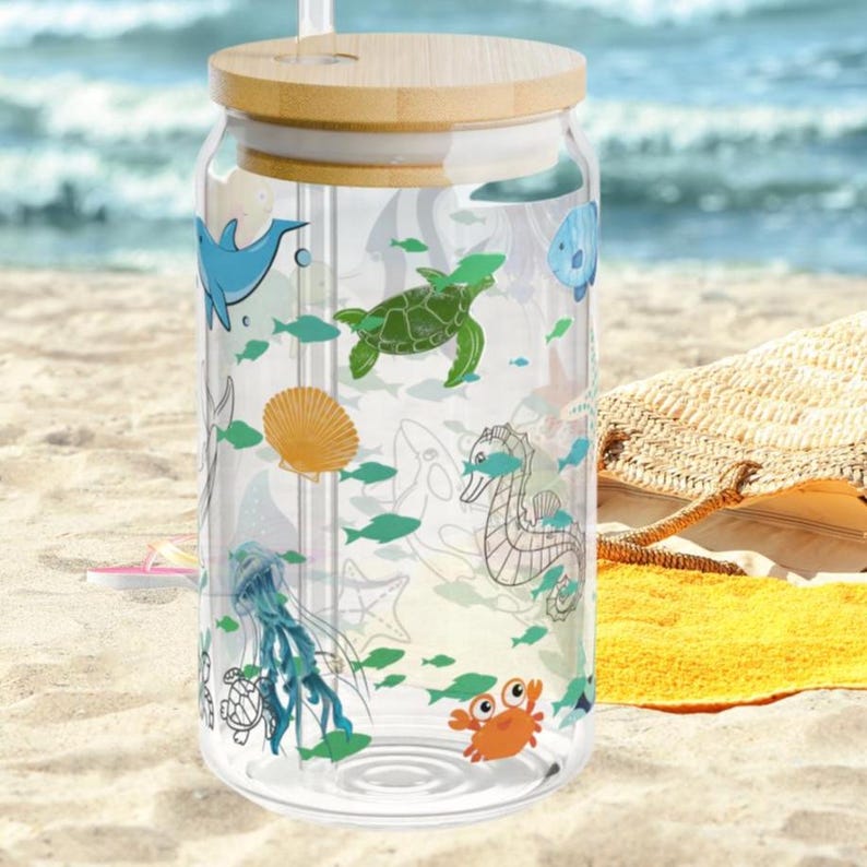 May include: A clear glass tumbler with a bamboo lid and a glass straw, featuring a colorful wrap-around print of various sea creatures including a blue dolphin, green sea turtle, orange crab, seahorse, jellyfish, and seashells.