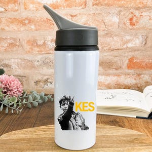 May include: White water bottle with a black and grey lid. The bottle has a black and white graphic of a person making a peace sign, with the word "KES" in yellow. The bottle is cylindrical.