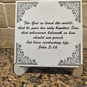 May include: A white square tile with black decorative corner designs and the text "For God so loved the world, that he gave his only begotten Son, that whosoever believeth in him should not perish but have everlasting life. John 3:16". The tile is displayed on a wooden stand.