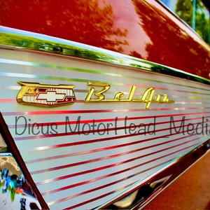 May include: Close-up of a red classic car with a chrome trim and a gold "Bel Air" emblem. The car has a white and red striped design on the side.