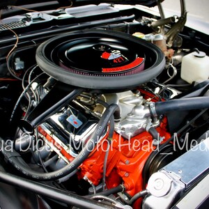 May include: Close-up of a classic car engine with a black air filter and red engine components. The engine is visible through the open bonnet of the car.