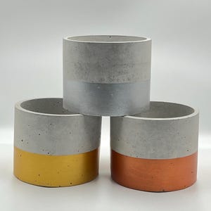 Puede incluir: Tres macetas de hormigón con un borde metálico. Las macetas son grises con un borde plateado, dorado y cobre.