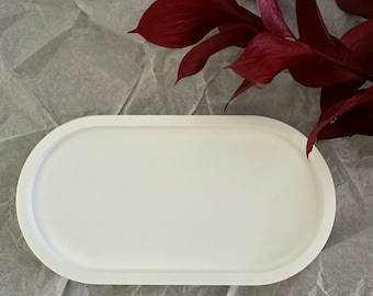 May include: A white, oval-shaped tray with a raised edge. The tray is made of a smooth material and sits on a textured, gray surface. Deep red leaves are in the background, adding a pop of color.
