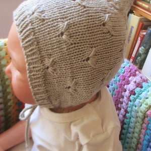 Könnte beinhalten: Ein Baby trägt eine beige gestrickte Mütze mit Rautenmuster. Die Mütze hat einen Bindfaden, der unter dem Kinn gebunden ist. Das Baby trägt ein weißes Hemd.