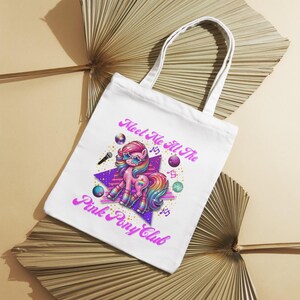 May include: White tote bag with a pink pony wearing sunglasses and a rainbow mane. The text on the bag reads "Meet Me At The Pink Pony Club".