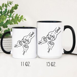 May include: Two white ceramic mugs with black handles and black rims. The mugs feature a black and white illustration of five fists with the words "Dad", "Isla", "Jaden", and "Dad" written on them. The mugs are 11 oz and 15 oz.