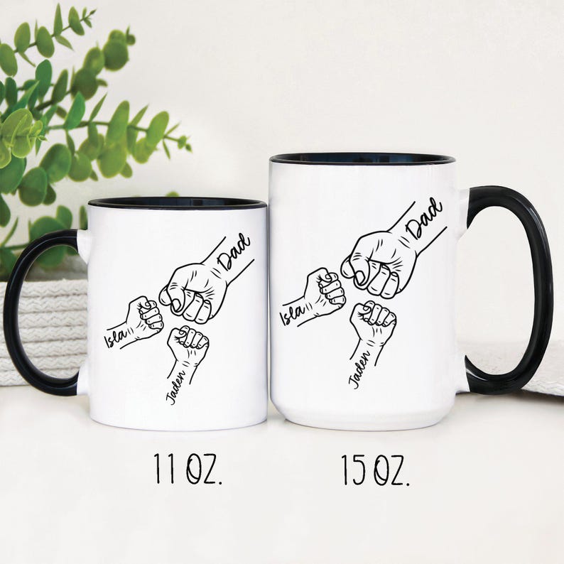 May include: Two white ceramic mugs with black handles and black rims. The mugs feature a black and white illustration of five fists with the words "Dad", "Isla", "Jaden", and "Dad" written on them. The mugs are 11 oz and 15 oz.