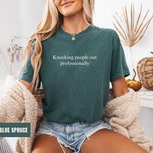 May include: A woman wearing a blue spruce t-shirt with the text "Knocking people out professionally".