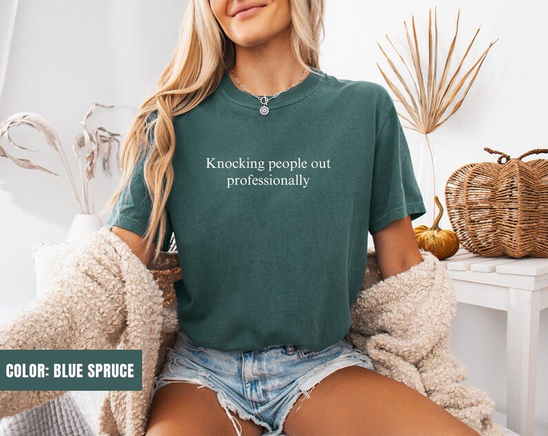 May include: A woman wearing a blue spruce t-shirt with the text "Knocking people out professionally".
