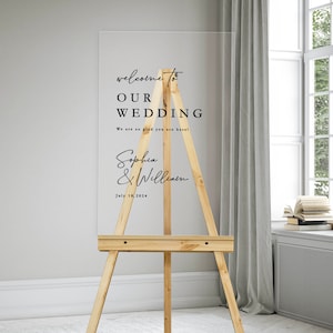 K&ouml;nnte beinhalten: Ein klares Acryl-Hochzeitschild mit schwarzem Text auf einem Staffelei. Das Schild lautet "Willkommen zu unserer Hochzeit. Wir freuen uns so, dass Sie da sind! Sophia & William. 18. Juli 2024."