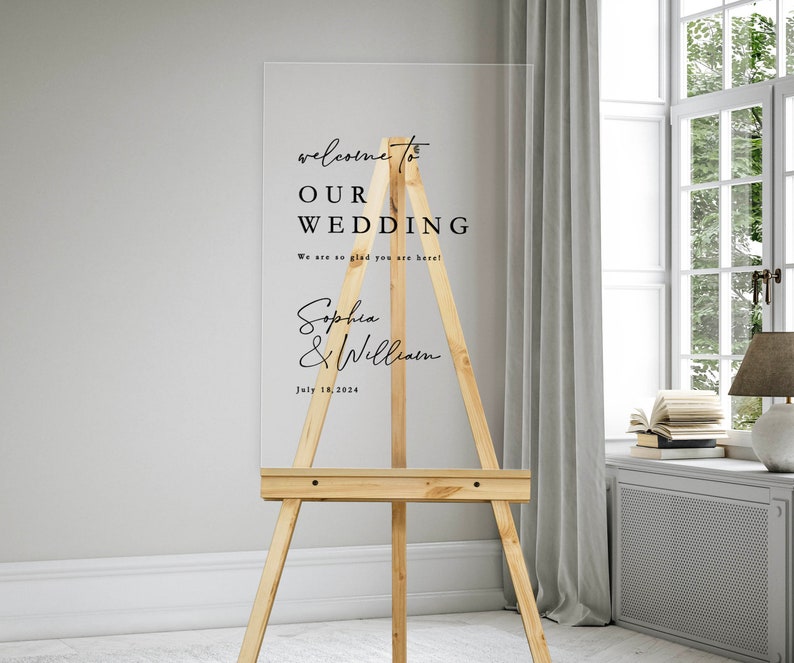 K&ouml;nnte beinhalten: Ein klares Acryl-Hochzeitschild mit schwarzem Text auf einem Staffelei. Das Schild lautet "Willkommen zu unserer Hochzeit. Wir freuen uns so, dass Sie da sind! Sophia & William. 18. Juli 2024."