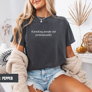 May include: A dark grey t-shirt with the text "Knocking people out professionally" printed on the front. The model is wearing the shirt with denim shorts.