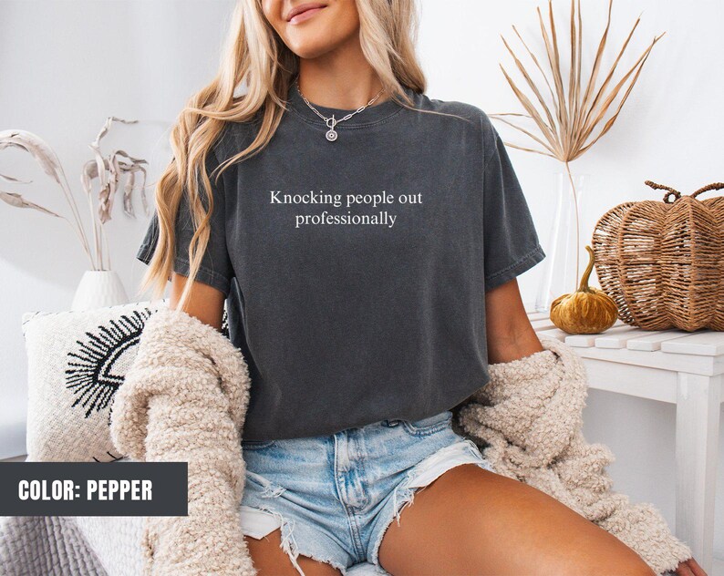 May include: A dark grey t-shirt with the text "Knocking people out professionally" printed on the front. The model is wearing the shirt with denim shorts.