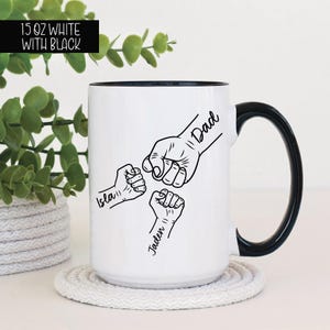 May include: White ceramic mug with a black handle. The mug has a black and white illustration of three fists with the names "Dad", "Isla", and "Jaden" written on them.