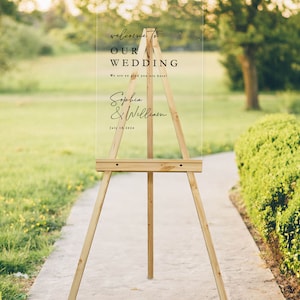 K&ouml;nnte beinhalten: Ein klares Acryl-Hochzeitschild mit schwarzem Text auf einem Staffelei. Das Schild lautet "Willkommen zu unserer Hochzeit" mit den Namen des Paares "Sophia & William" und dem Datum "18. Juli 2024".