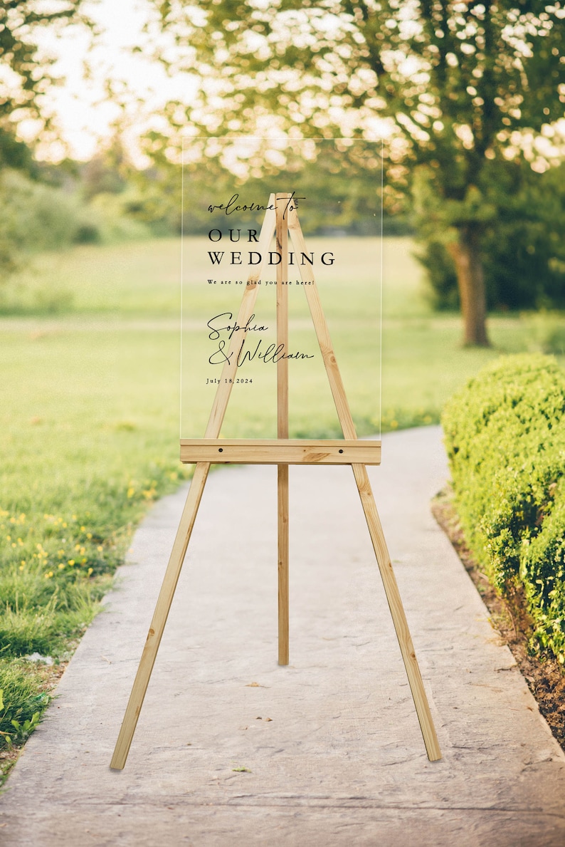 K&ouml;nnte beinhalten: Ein klares Acryl-Hochzeitschild mit schwarzem Text auf einem Staffelei. Das Schild lautet "Willkommen zu unserer Hochzeit" mit den Namen des Paares "Sophia & William" und dem Datum "18. Juli 2024".