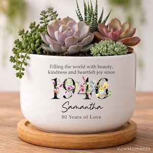 May include: A white ceramic planter filled with succulents, resting on a wooden coaster. The planter has the text "Filling the world with beauty, kindness and heartfelt joy since 1946 Samantha 80 Years of Love."