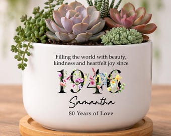May include: A white ceramic planter filled with succulents, resting on a wooden coaster. The planter has the text "Filling the world with beauty, kindness and heartfelt joy since 1946 Samantha 80 Years of Love."