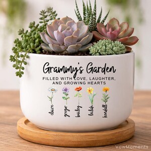 May include: A white ceramic planter with the text "Grammy's Garden" and the words "Filled with Love, Laughter, and Growing Hearts." The planter contains various succulents and is set on a wooden base.