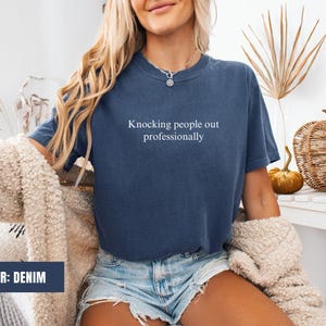 May include: A denim blue t-shirt with the text "Knocking people out professionally" printed in white on the front.