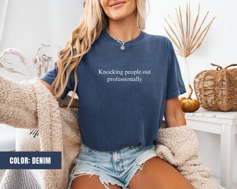 May include: A denim blue t-shirt with the text "Knocking people out professionally" printed in white on the front.