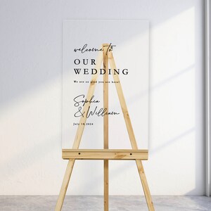 K&ouml;nnte beinhalten: Ein wei&szlig;es Acryl-Hochzeitschild mit schwarzem Text auf einem Staffelei. Das Schild lautet "Willkommen zu unserer Hochzeit. Wir freuen uns so, dass Sie da sind! Sophia & William. 18. Juli 2024."