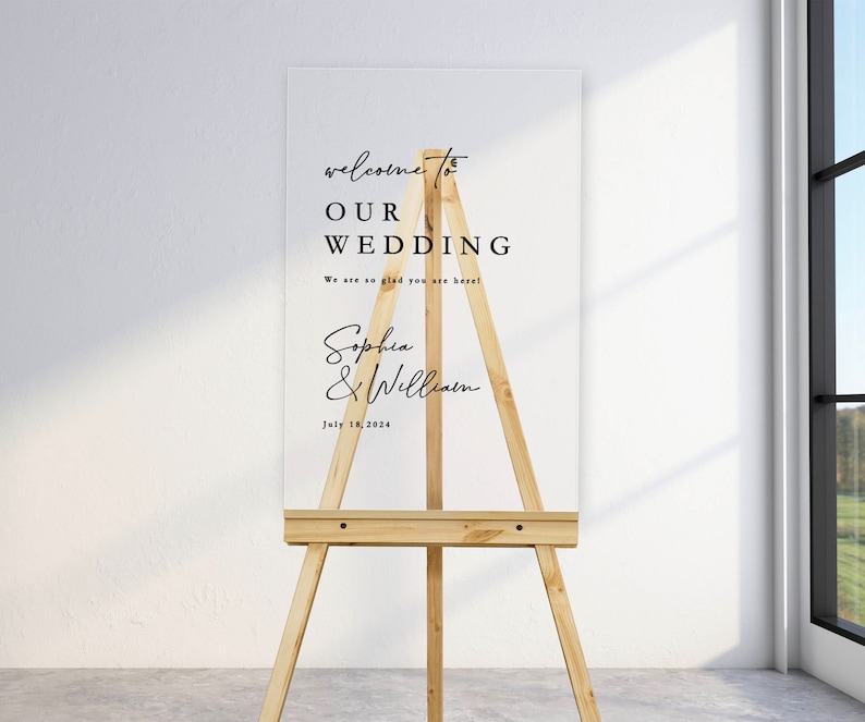 K&ouml;nnte beinhalten: Ein wei&szlig;es Acryl-Hochzeitschild mit schwarzem Text auf einem Staffelei. Das Schild lautet "Willkommen zu unserer Hochzeit. Wir freuen uns so, dass Sie da sind! Sophia & William. 18. Juli 2024."