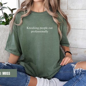 May include: A moss green t-shirt with the text "Knocking people out professionally" printed in white on the front. The shirt is being worn by a person with long brown hair and blue jeans.